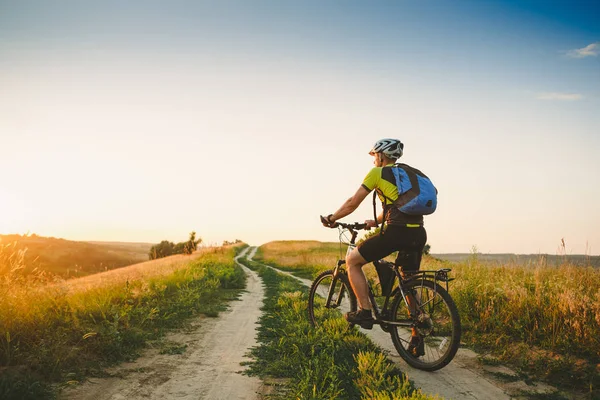 Joven ciclismo en un camino rural a través del prado de verano puesta de sol — Foto de Stock