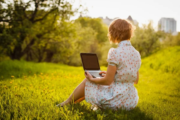 Молодая женщина с ноутбуком сидит на зеленой траве в парке на закате — стоковое фото