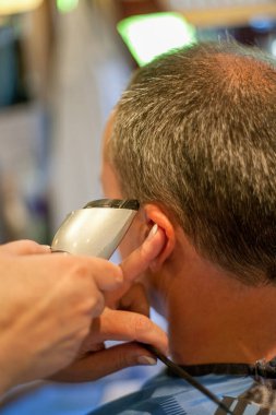 Kuaför, saç kesme makinesiyle kafasını tıraş ediyor ya da saç makası kullanıyor. Müşterinin arkasından görülüyor..