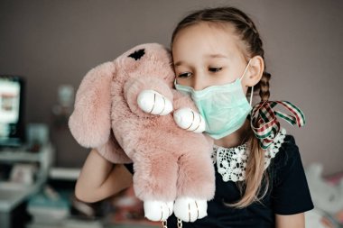 Tıbbi maskeli küçük bir kız oyuncak tutuyor, Corona virüsünden korunuyor.