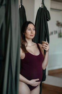 Hamile bir kadın stüdyoda yoga yapıyor. Sıkı vücut giyiyor. Balyasını kucaklıyor ve gelecekteki bebeğine çok dikkat ediyor. Anne bakımı kavramı, genç anne, kadın ve çocuk, sağlıklı yaşam, fitness. 