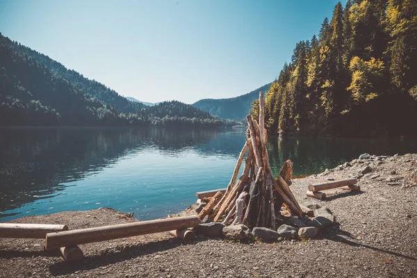 Prepared for kindling a large bonfire and benches from the logs on the shore of a beautiful lake with clear water surrounded by mountains and forest. Toned image