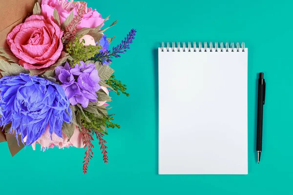 Artificial handmade flowers, a notebook and a black pen on a turquoise background