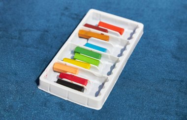 Beyaz bir kutunun içindeki renkli boya kalemleri mavi bir yüzeyde yatıyor.