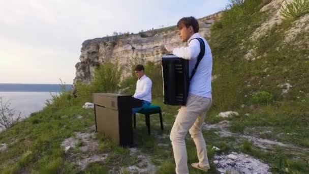 Два музыканта Пианист и аккордеонист играют на скалах на закате над морем — стоковое видео