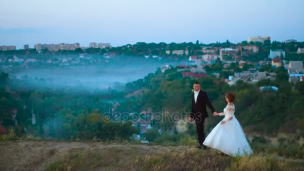 Pareja joven enamorada en un prado de montaña en el fondo de la niebla y la ciudad 4k — Vídeos de Stock