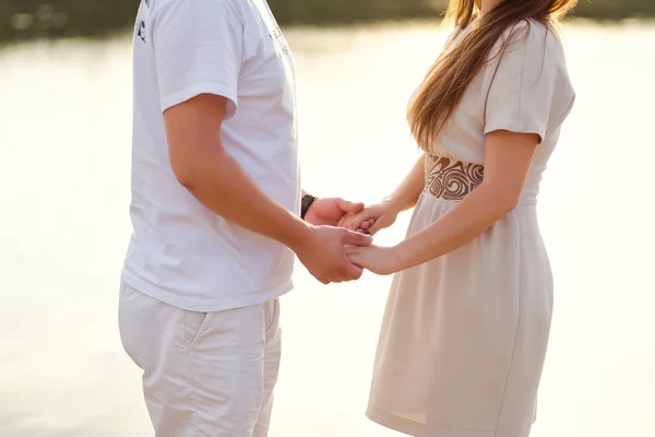 Ungt par hålla händerna på stranden av floden, händer närbild, kärlek, känslor, solnedgång vid floden, bukett prästkragar — Stockfoto