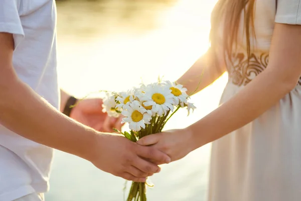 Ungt par hålla händerna på stranden av floden, händer närbild, kärlek, känslor, solnedgång vid floden, bukett prästkragar — Stockfoto