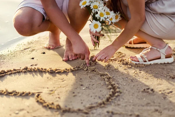 Pasangan muda jatuh cinta menggambar hati di atas pasir, pertemuan pasangan muda, hati di pasir, tangan close-up buket aster, matahari terbenam di dekat air, malam musim panas — Stok Foto