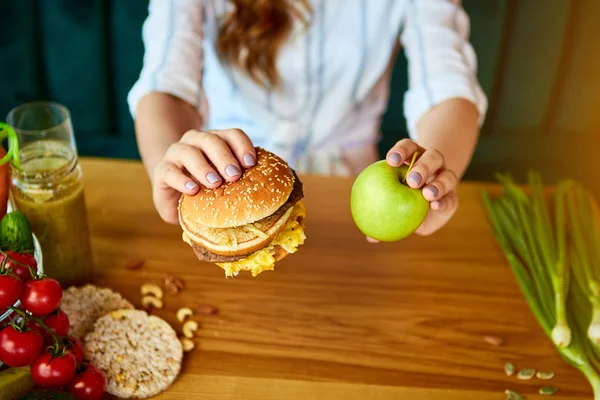 Όμορφη νεαρή γυναίκα αποφασίζει να φάει χάμπουργκερ ή μήλο στην κουζίνα. Φτηνές πρόχειρο φαγητό εναντίον υγιεινής διατροφής — Φωτογραφία Αρχείου