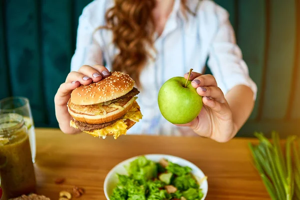 美しい若い女性は台所でハンバーガーやリンゴを食べることを決定します.安いジャンクフード対健康的な食事 — ストック写真