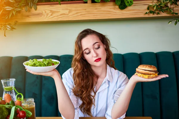 Όμορφη νεαρή γυναίκα αποφασίζει να φάει χάμπουργκερ ή φρέσκια σαλάτα στην κουζίνα. Φτηνές πρόχειρο φαγητό εναντίον υγιεινής διατροφής — Φωτογραφία Αρχείου