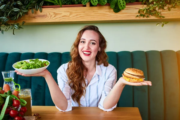 美しい若い女性はキッチンでハンバーガーや新鮮なサラダを食べることを決定します.安いジャンクフード対健康的な食事 — ストック写真