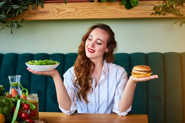 美しい若い女性はキッチンでハンバーガーや新鮮なサラダを食べることを決定します.安いジャンクフード対健康的な食事 — ストック写真