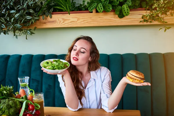 Красивая молодая женщина решает есть гамбургер или свежий салат на кухне. Дешевая вредная пища против здорового питания — стоковое фото