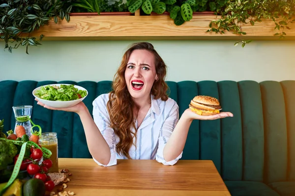 美しい若い女性はキッチンでハンバーガーや新鮮なサラダを食べることを決定します.安いジャンクフード対健康的な食事 — ストック写真