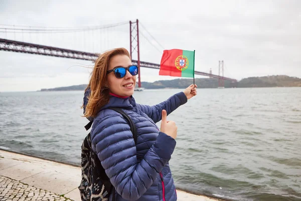 Ung kvinna turist som innehar Portugals flagga i händerna och njuter av landskaps syn på den berömda Iron Bridge 25 april stående tillbaka på Riverside i Lissabon City — Stockfoto