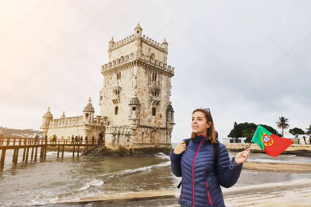 Young woman tourist walking near Belem tower holding the flag of Portugal in hands on the riverside in Lisbon