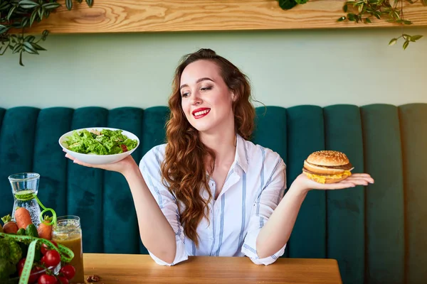 Красивая молодая женщина решает есть гамбургер или свежий салат в — стоковое фото