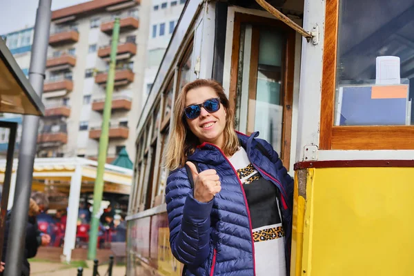 葡萄牙里斯本市大街上著名的复古黄色电车旁，站着一位年轻而快乐的女游客 — 图库照片