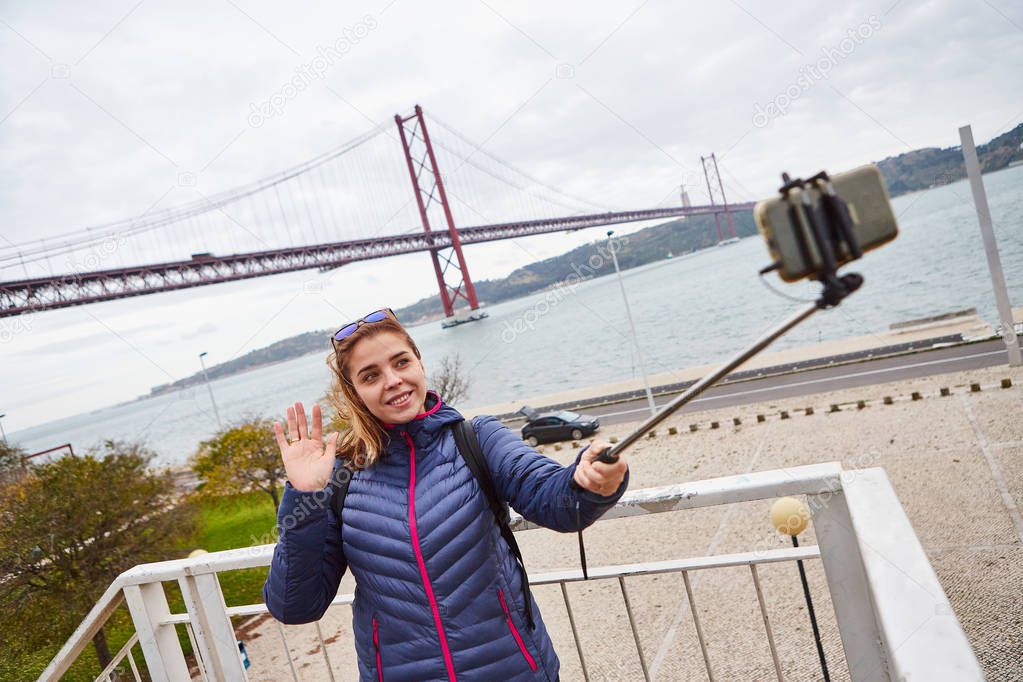 Young woman tourist take selfies on the background of famous iron 25th of April bridge in Lisbon city, Portugal