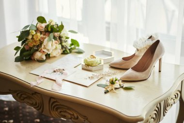 Ayakkabı, buket, yüzük ve davetiye kartları gibi düğün aksesuarları masanın üzerinde.