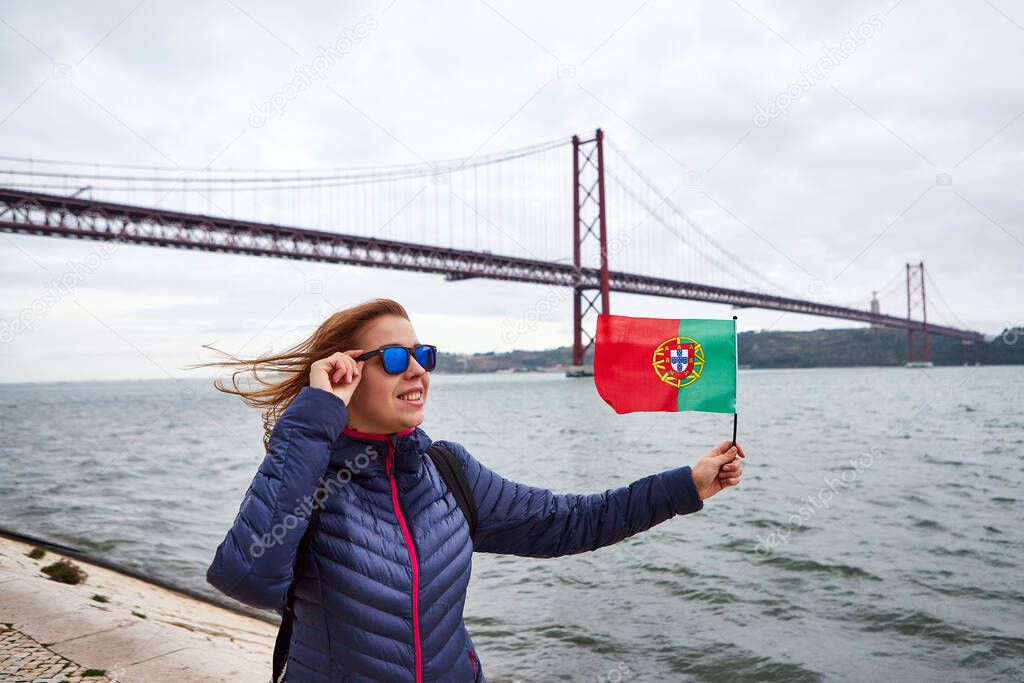 Young woman tourist holding the flag of Portugal in hands and enjoying landscape view on the famous iron bridge 25th of April standing back on the riverside in Lisbon city