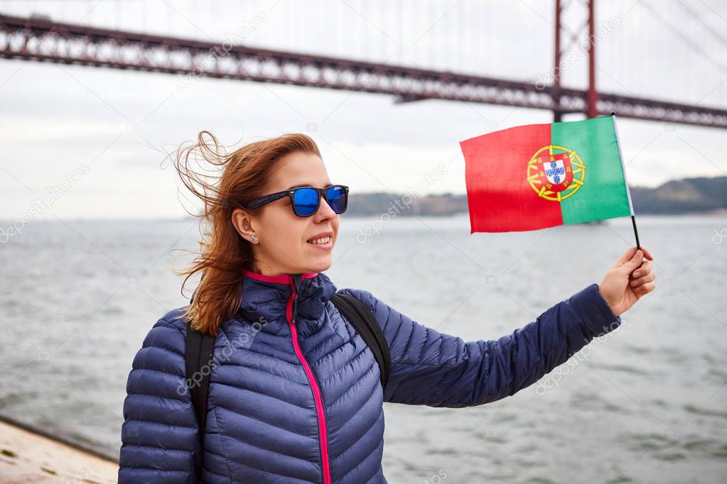 Young woman tourist holding the flag of Portugal in hands and enjoying landscape view on the famous iron bridge 25th of April standing back on the riverside in Lisbon city