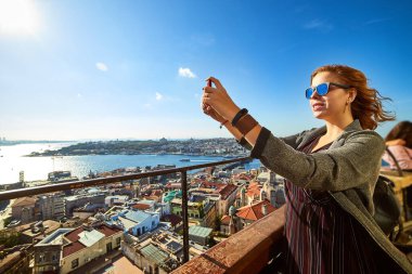 Güzel bir turist kız akıllı telefonuyla fotoğraf çekiyor ve Galata Kulesi 'nin tepesinden İstanbul' un tarihi merkezinin manzarasını seyrediyor.