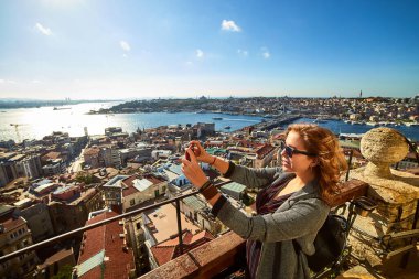 Güzel bir turist kız akıllı telefonuyla fotoğraf çekiyor ve Galata Kulesi 'nin tepesinden İstanbul' un tarihi merkezinin manzarasını seyrediyor.