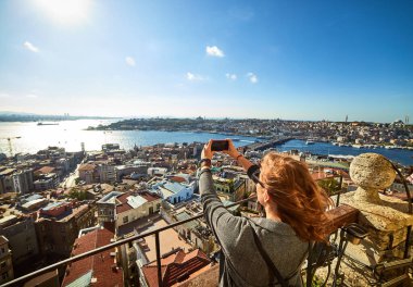 Güzel bir turist kız akıllı telefonuyla fotoğraf çekiyor ve Galata Kulesi 'nin tepesinden İstanbul' un tarihi merkezinin manzarasını seyrediyor.