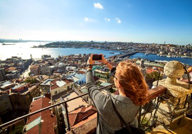 Güzel bir turist kız akıllı telefonuyla fotoğraf çekiyor ve Galata Kulesi 'nin tepesinden İstanbul' un tarihi merkezinin manzarasını seyrediyor.