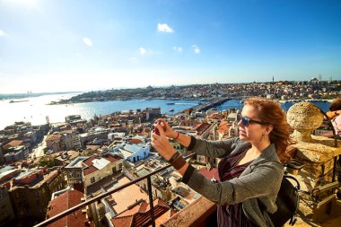 Güzel bir turist kız akıllı telefonuyla fotoğraf çekiyor ve Galata Kulesi 'nin tepesinden İstanbul' un tarihi merkezinin manzarasını seyrediyor.