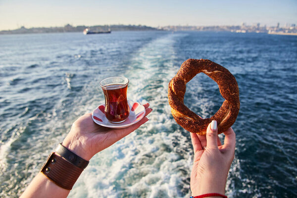 Чашка (броненосец) традиционного турецкого чая и симитчи (круглый бублик с семенами кунжута) в руке на фоне городского пейзажа Стамбула и моря Мармар
