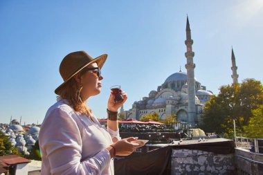 Şapkalı mutlu çekici kadın turist, İstanbul 'daki Süleyman Camii' nin arka planında geleneksel Türk çayı (armuda) içiyor. Din ve seyahat kavramı.