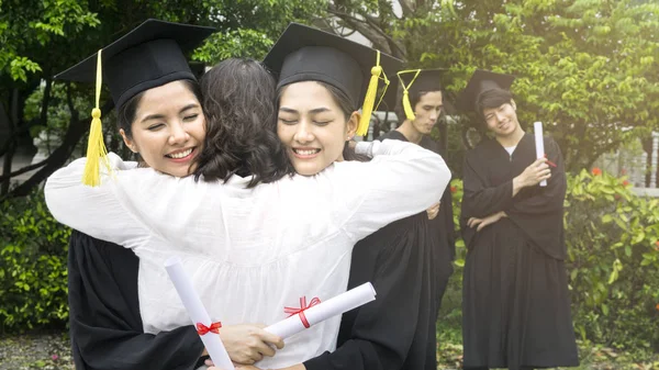 Due studentesse con gli abiti di laurea e cappello abbracciare il genitore in cerimonia di congratulazione con gli studenti ragazzo sullo sfondo . — Foto Stock
