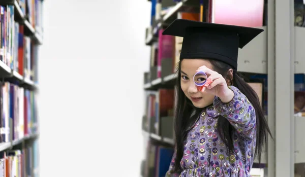 La piccola ragazza carina con il cappello di laurea e il diploma di carta stand e sorridere nello scaffale dei libri di sfondo . — Foto Stock