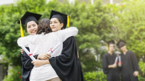 Dos asiático chica estudiantes con la graduación batas y sombrero abrazo el padre en felicitación ceremonia con desenfoque de chico estudiantes en fondo . — Foto de Stock