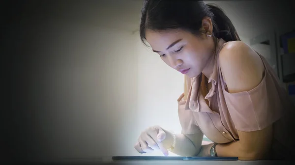 Ásia adolescente mulher lê e toca no um tablet dispositivo no escuro luz processo fundo — Fotografia de Stock