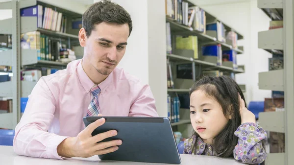 Hombre profesor y niño estudiante aprendizaje y mirando en el dispositivo de la tableta con el libro del uno mismo en el fondo — Foto de Stock