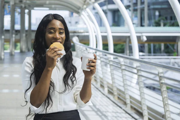 Portrait of happy business black woman eats on hold fast food hamburger and cup of coffee in the outdoor pedestrian walk way with the city space background