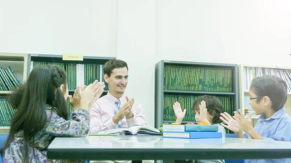 Sonriente caucásico maestro y agrupación de asiático niños estudiante aprendizaje y hablar en blanco mesa y color libro con librería fondo — Foto de Stock
