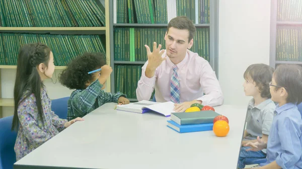 Sonriente caucásico maestro y agrupación de asiático niños estudiante aprendizaje y hablar en blanco mesa y color libro con librería fondo — Foto de Stock