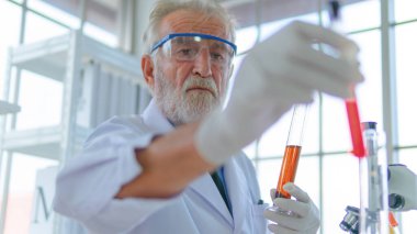 Kıdemli profesör erkek araştırmacı, laboratuvar bilimi için yüz konsantrasyonuyla kimyasal sıvı tüpü test ediyor. Beyaz labaratuvarı ve masasında ekipmanları var..