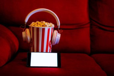 Kırmızı koltukta patlamış mısır kovası olan boş beyaz ekranlı cep telefonu. İnternet telefonunda TV yayını kavramı.
