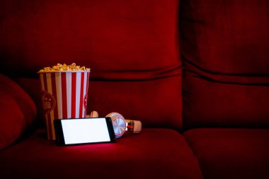 Kırmızı koltukta patlamış mısır kovası olan boş beyaz ekranlı cep telefonu. İnternet telefonunda TV yayını kavramı.
