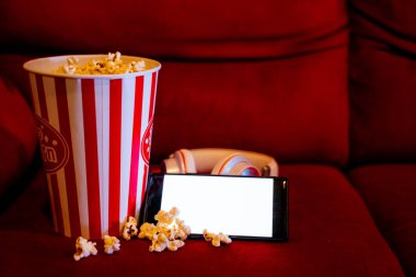 Kırmızı koltukta patlamış mısır kovası olan boş beyaz ekranlı cep telefonu. İnternet telefonunda TV yayını kavramı.