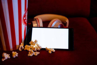 Kırmızı koltukta patlamış mısır kovası olan boş beyaz ekranlı cep telefonu. İnternet telefonunda TV yayını kavramı.