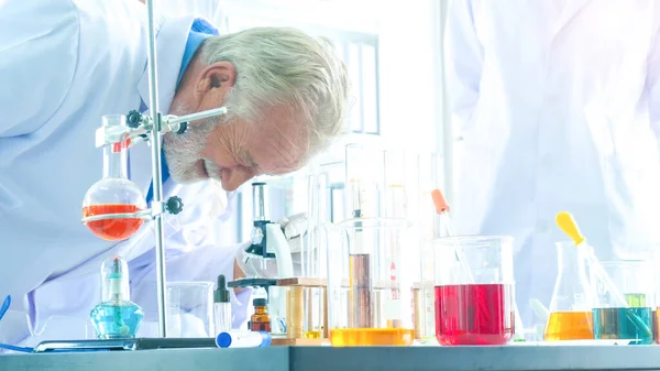 close up face of Senior male scientist seeing microscope. Man Research is working in the Laboratory. With foreground of tube and Beaker of liquid substance.
