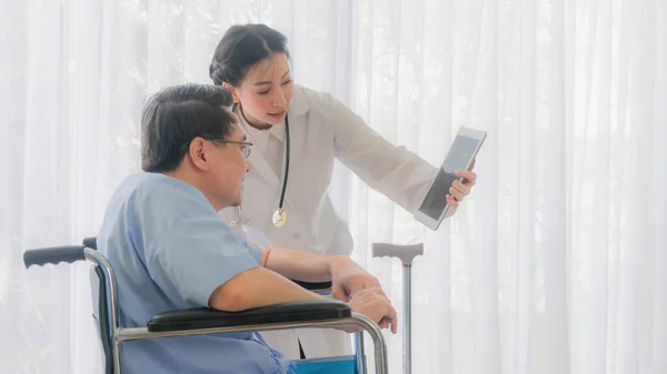 Asian woman doctor gives advice on tablet computer to an elderly old senior patient sit in wheelchair in Interior patient room background white curtain in hospital.